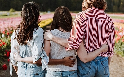 Three people stand in a field of flowers, facing away from the camera with their arms wrapped around each other.