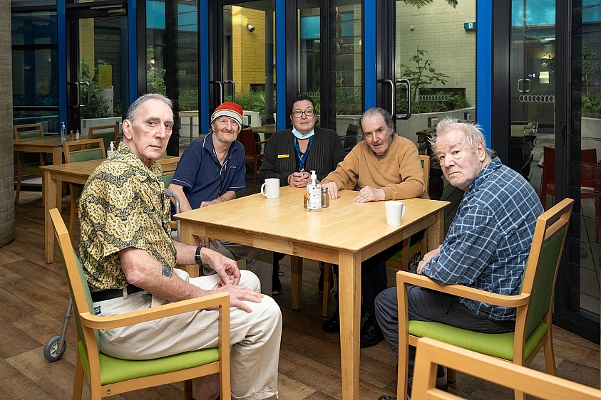 A group of people sitting around a kitchen table, some drinking tea and coffee.