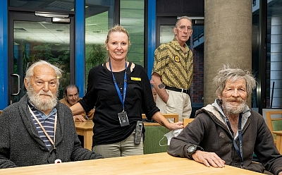 Two men sit at a table, with a person wearing a lanyard standing behind them, resting their hand on the men's shoulders.